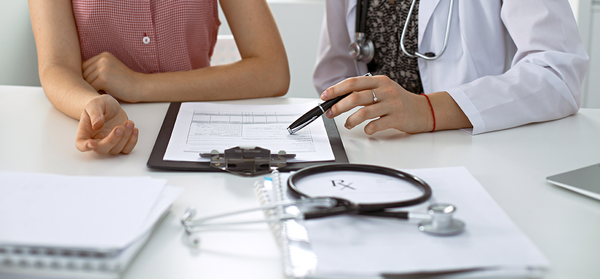 A doctor and patient sit at a table and are reviewing a document on a clipboard. In the foreground some of the doctors medical tools can be seen.