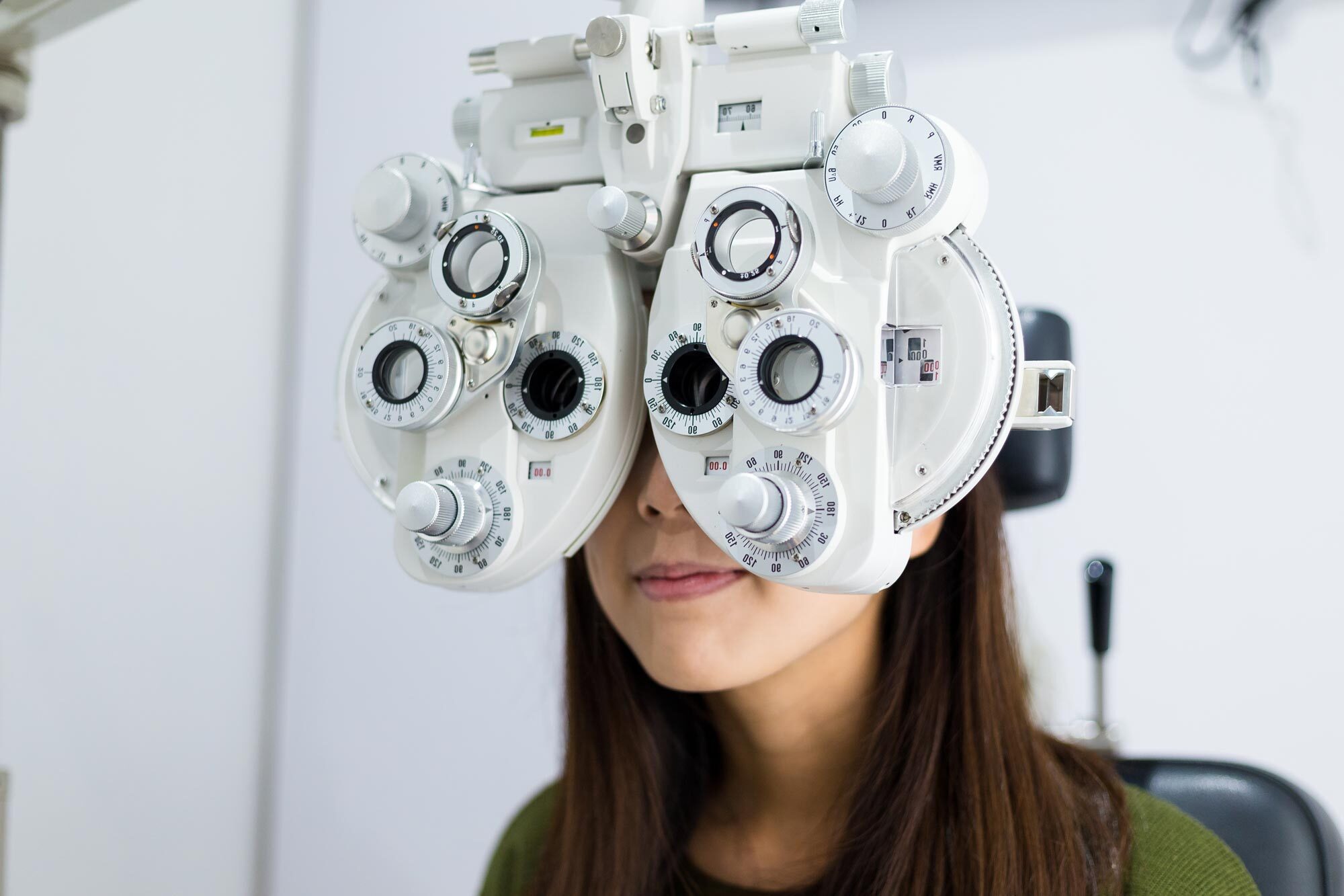 A woman with brown hair is sitting in an exam chair while looking through a white phoropter machine.