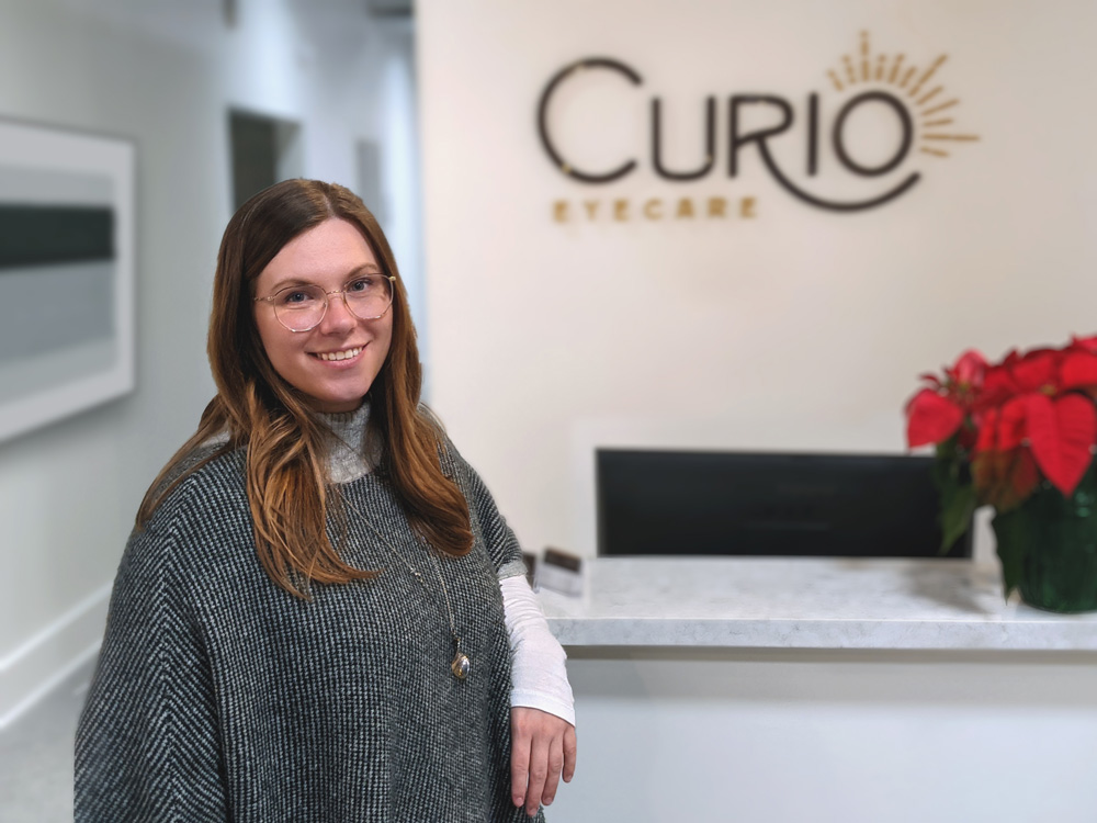 Dr. Lorencz is posed infront of her reception desk. She is smiling at the camera while the logo of Curio eyecare can be seeing blurred in the background.