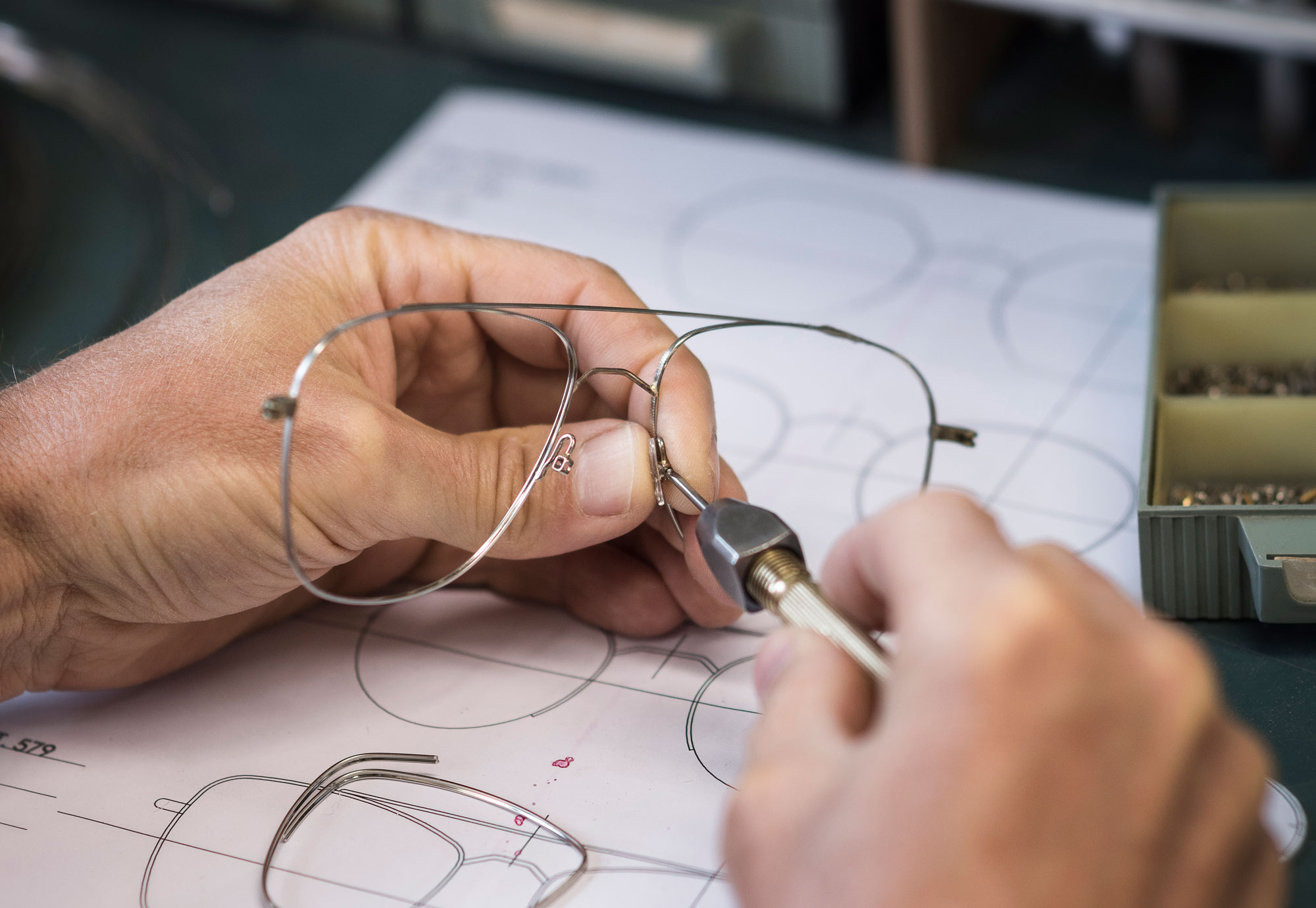 A man tightly holds a pair of metal glasses. He appears to be at a work station and is using a tool to manipulate the front of the glasses.
